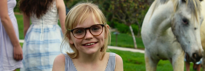 A young girl smiling in glasses. There is a horse behind her.